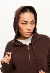 Sudadera con cremallera marrón, con bordes acanalados, que presenta una capucha con cordón y una textura acogedora. El modelo sostiene el cordón cerca del cierre de la cremallera.