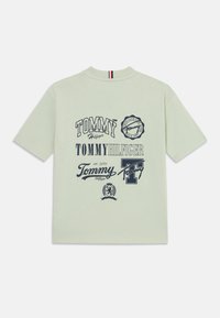 Lys grønn t-skjorte med korte ermer som har ulike marineblå logoer og teksttrykk, inkludert "Tommy Hilfiger" og designaksenter. Rund hals.