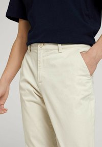 Beżowe bawełniane spodnie o prostym kroju, z przednimi kieszeniami i zapinanym na guziki paskiem, w połączeniu z ciemnogranatową koszulą z krótkim rękawem.