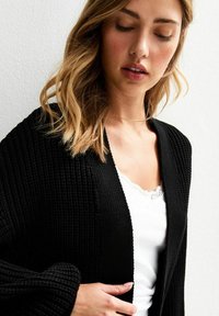 Cardigan nero con texture, maniche lunghe e design aperto davanti. Indossato sopra una canottiera bianca con pizzo.