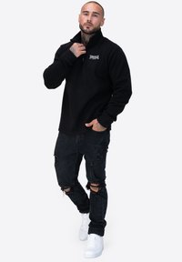 Czarny polar z ćwierć zamkiem i kołnierzem, w połączeniu z przetartymi czarnymi dżinsami i białymi tenisówkami, z logo na piersi.