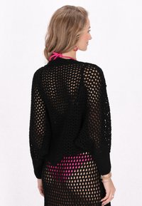 Czarny sweter o otwartym splotem z długimi rękawami, o luźnym kroju i dużymi wzorami w romby. Nosi się go na różowy strój kąpielowy, podkreślając przezroczystość.