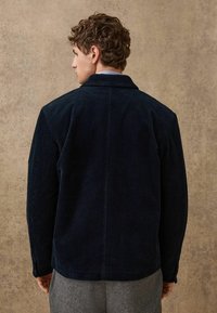 Giacca in corduroy navy con colletto classico e maniche lunghe, caratterizzata da una texture a coste verticali. La vista posteriore mostra una vestibilità sartoriale, con dettagli minimali.
