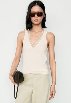 Donna con capelli neri lisci che indossa occhiali da sole marroni, un top beige senza maniche con rifinitura in pizzo, pantaloni chiari e tiene una piccola borsa marrone scuro.