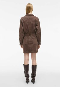 Smeđi traper haljina s dugim rukavima, remenom u struku i dvjema stražnjim džepovima; sadrži detalje prošivanja i opušteni kroj. Dužine do koljena, jednostavan dizajn.