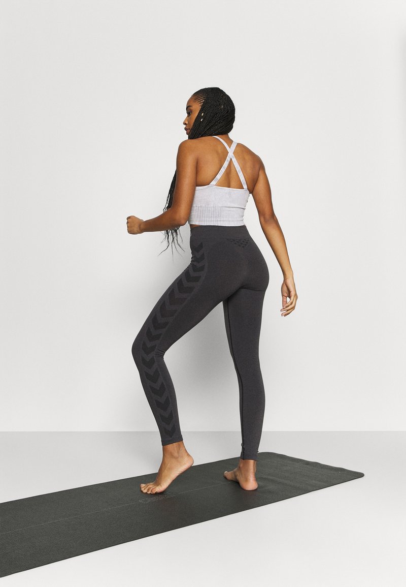 Mujer con trenzas largas que lleva una camiseta deportiva blanca con tirantes cruzados y leggings negros con diseño de chevrón, de pie descalza sobre una esterilla negra de yoga.