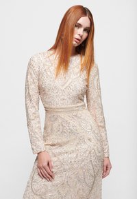 Perlenbesetztes Kleid mit langen Ärmeln in Creme, mit filigranen Wellenmustern und silbernen Akzenten. Es hat eine taillierte Silhouette und einen runden Ausschnitt.