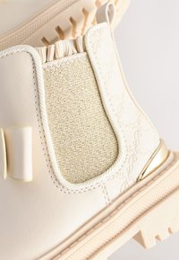 Bottes blanches montant à la cheville avec un panneau élastique texturé doré, des coutures décoratives et une semelle en caoutchouc avec un motif de crampons épais.