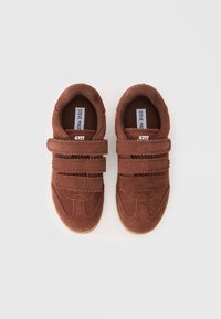 Sapatos de camurça castanhos com duas tiras de Velcro, biqueira arredondada e sola de borracha. Apresenta um acabamento em zigzag nas tiras e uma superfície texturizada.
