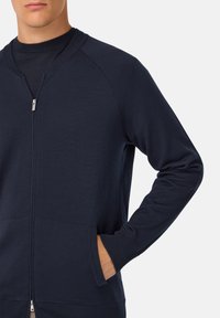 Mornarska zip majica s kapuco z dolgimi rokavi, ribastimi manšetami in dvema sprednjima žepoma. Narejena iz mehkega, teksturiranega materiala s preprostim dizajnom.