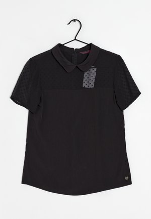 Sort kortærmet bluse med tekstureret prikket gennemsigtigt øverste del, Peter Pan-krave, lynlås bagpå og mærke på en sort bøjle mod hvid baggrund.