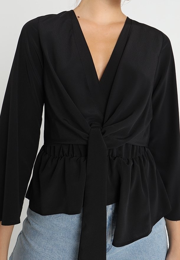 Blouse noire à manches longues avec un col en V profond et un détail noué devant, portée avec un jean bleu clair sur une personne des épaules aux hanches.