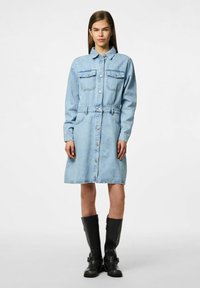 Light blue denim shirt dress with long sleeves, button front, two chest pockets, drawstring waist, and a flared skirt. Black knee-high boots.