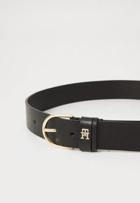 Ceinture en cuir noir avec une boucle en métal doré. Elle présente des accessoires en or et un petit logo sur la sangle en cuir.