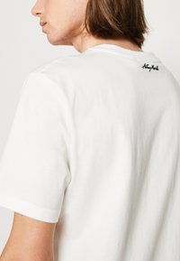 Camiseta de algodón blanca con mangas cortas, con un cuello redondo y un logo bordado negro en la parte superior de la espalda. Textura suave.