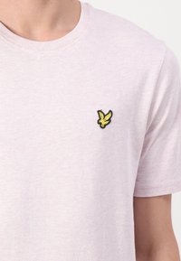 T-shirt en coton rose clair avec un col rond et des manches courtes. Présente un petit logo de bird noir et jaune sur la partie gauche de la poitrine.