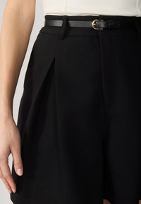 Pantaloni corti neri su misura realizzati in tessuto intrecciato, con pieghe, vita aderente e una cintura nera sottile con una fibbia dorata. Texture liscia.