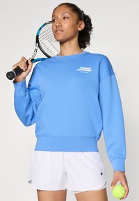 Sweatshirt de algodão azul claro com punhos e bainha canelados, apresentando texto branco que diz "TÊNIS ALGUÉM? BJÖRN BORG." Usado com calções brancos, segurando uma bola de ténis e uma raquete.
