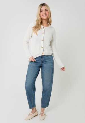 Mujer rubia sonriendo, vistiendo un cárdigan blanco de punto con botones y bolsillos, jeans azules de talle alto y zapatos planos beige, de pie sobre un fondo blanco.