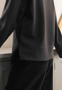 Schwarzer Pullover mit langen Ärmeln, gerippter Struktur, mit schrägen Seitennähten und leicht kurzem Schnitt, kombiniert mit schwarzen Cordhosen.