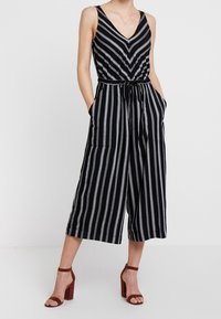 Combinaison rayée en noir et blanc avec un décolleté en V profond, taille cintrée, jambes larges et cropped, et poches latérales. Associée à des sandales à talons.