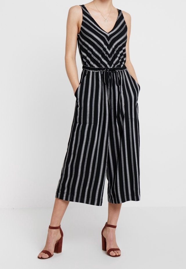 Combinaison rayée en noir et blanc avec un décolleté en V profond, taille cintrée, jambes larges et cropped, et poches latérales. Associée à des sandales à talons.