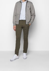 Grå zip-up tröja, randig skjorta med knappar, olivgröna byxor och vita sneakers. Tröjan har en krage och ribbade muddar.