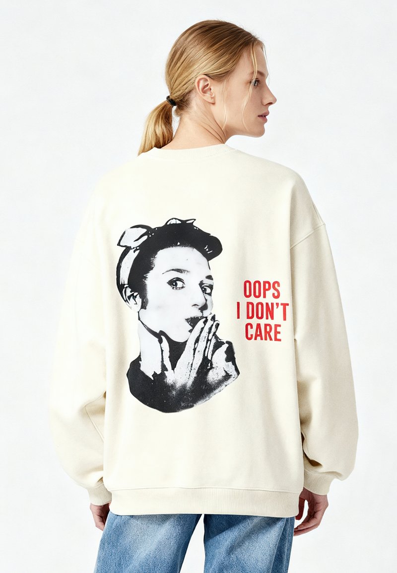 Krēmkrāsas pārizmērīga džemperis ar melnbaltu grafiku, kurā attēlota sieviete ar loku, un sarkaniem burtiem uz muguras uzraksts „OOPS I DON'T CARE”.