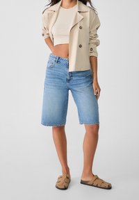 Veste cropped beige clair, haut ajusté crème, et bermuda en denim bleu moyen. La personne porte des sandales à glisser brunes.