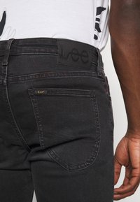 Närbild av en person som bär svarta Lee-jeans med synlig bakficka och en svart läder Lee-märke på midjebandet.