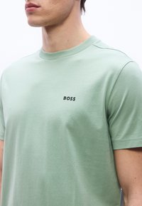 Κοντινό πλάνο ενός ατόμου που φοράει ένα ανοιχτό πράσινο μπλουζάκι με μικρό μαύρο λογότυπο "BOSS" στο αριστερό στήθος.