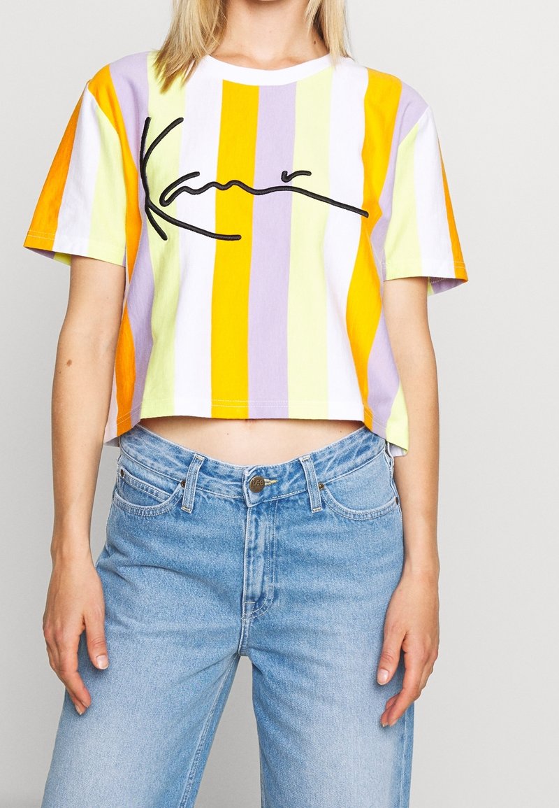 Lyhythihainen t-paita, jossa on pystysuoria raitoja oranssina, keltaisena, purppuran värisenä ja valkoisena. Keskellä on musta kaunis logo. Yhdistetty vaaleansinisiin farkkuihin.