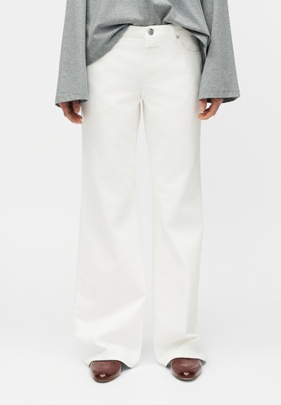Personne portant un pantalon blanc à jambes larges, un haut gris à manches longues et des chaussures marron à bout pointu, debout devant un fond uni.