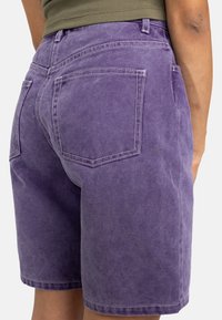 Shorts in misto cotone viola con una vestibilità comoda, cuciture a vista e due tasche posteriori. Il tessuto ha una leggera trama e un aspetto sbiadito.