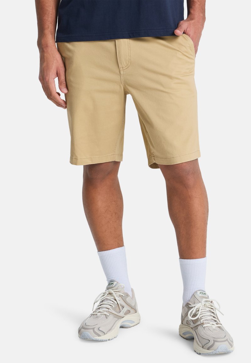 Bermuda beige realizzati in tessuto leggero, con una vestibilità classica, tasche anteriori e dettagli cuciti. Indossati con sneaker grigie e calzini bianchi.