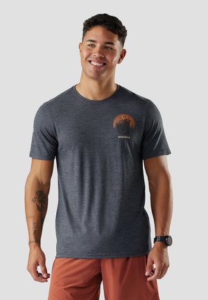 Glimlachende man die een donkergrijs T-shirt met print en oranje korte broek draagt, met tatoeages op zijn linkerarm en een zwart horloge om zijn rechterpols.
