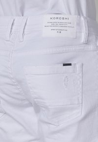 Pantalones de mezclilla blanca con bolsillos traseros, un ligero desgaste y una etiqueta con "KOROSHI". Las costuras planas y los lazos para el cinturón acentúan el diseño.