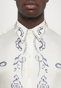 Nahaufnahme einer Person, die ein cremefarbiges Hemd mit schwarzer floralem Stickerei am Kragen und an der Vorderseite trägt, bis zum Hals zugeknöpft.