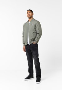 Giacca bomber verde con zip frontale e polsini a coste, abbinata a jeans in denim neri e sneakers nere su uno sfondo neutro.