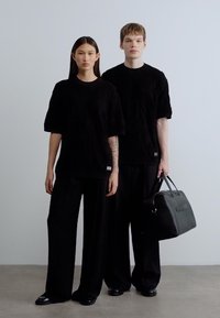 Μαύρα πλεκτά υπερμεγέθη τοπ, παντελόνια με φαρδύ πόδι και μαύρη τσάντα χειρός. Λιτές υφές και μινιμαλιστικός σχεδιασμός με διακριτικές λεπτομέρειες branding.