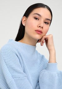Lyseblå strikket sweater med høj hals, der har en ribbet tekstur og flared ærmekanter. Modellen hviler sin hånd på sin hage.