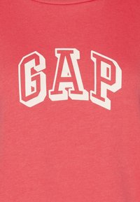 El sudadera color coral presenta un atrevido texto blanco "GAP" en una fuente gruesa. El material parece suave con una textura lisa y un diseño estándar de cuello redondo.