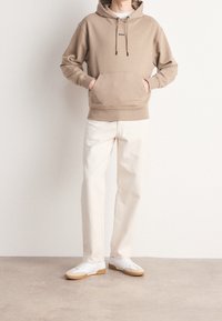 Sudadera beige con un bolsillo frontal, cordones y logo en el pecho, combinada con jeans de color claro y zapatillas blancas.