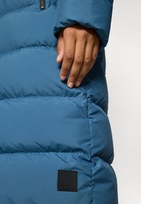 Blue puffer jacket with a smooth, shiny texture. Features a black logo patch and a hand resting on the side seam.