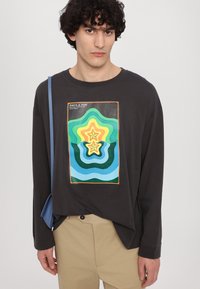 Čierne tričko s dlhým rukávom so farebnou vlnitou grafikou na prednej strane. V kombinácii s béžovými nohavicami a modrou taškou. Hladká textúra.