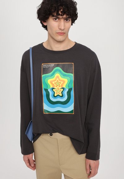 Zwart langemouw t-shirt met een kleurrijke golvende graphic op de voorkant. Gecombineerd met beige broeken en een blauwe tas. Glad textuur.