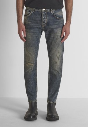 Man draagt vervaagde, versleten blauwe jeans en zwarte enkellaarsjes en staat tegen een lichte achtergrond.