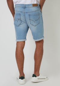 Pantalones cortos de mezclilla azul claro con puños enrollados, que presentan dos bolsillos traseros con costura curva y un parche de cuero marrón en la cintura.