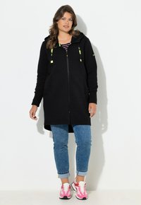 Sudadera negra con cremallera y capucha, de tela texturizada y cordones verdes, combinada con vaqueros azules remangados y zapatillas rosas.