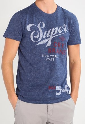 Mann trägt blaues T-Shirt mit der Aufschrift „Super Dry Brand New York State“ und hellgraue Shorts, Hände in den Taschen, vor schlicht weißem Hintergrund.